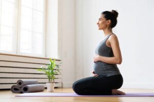 Una donna incinta in tenuta atletica si inginocchia su un tappetino da yoga all'interno, abbracciando il suo pancione e guardando fuori da una finestra luminosa, cogliendo l'essenza dello Yoga in gravidanza per il benessere di mamma bambino. Accanto a lei ci sono tappetini da yoga e una pianta in vaso.